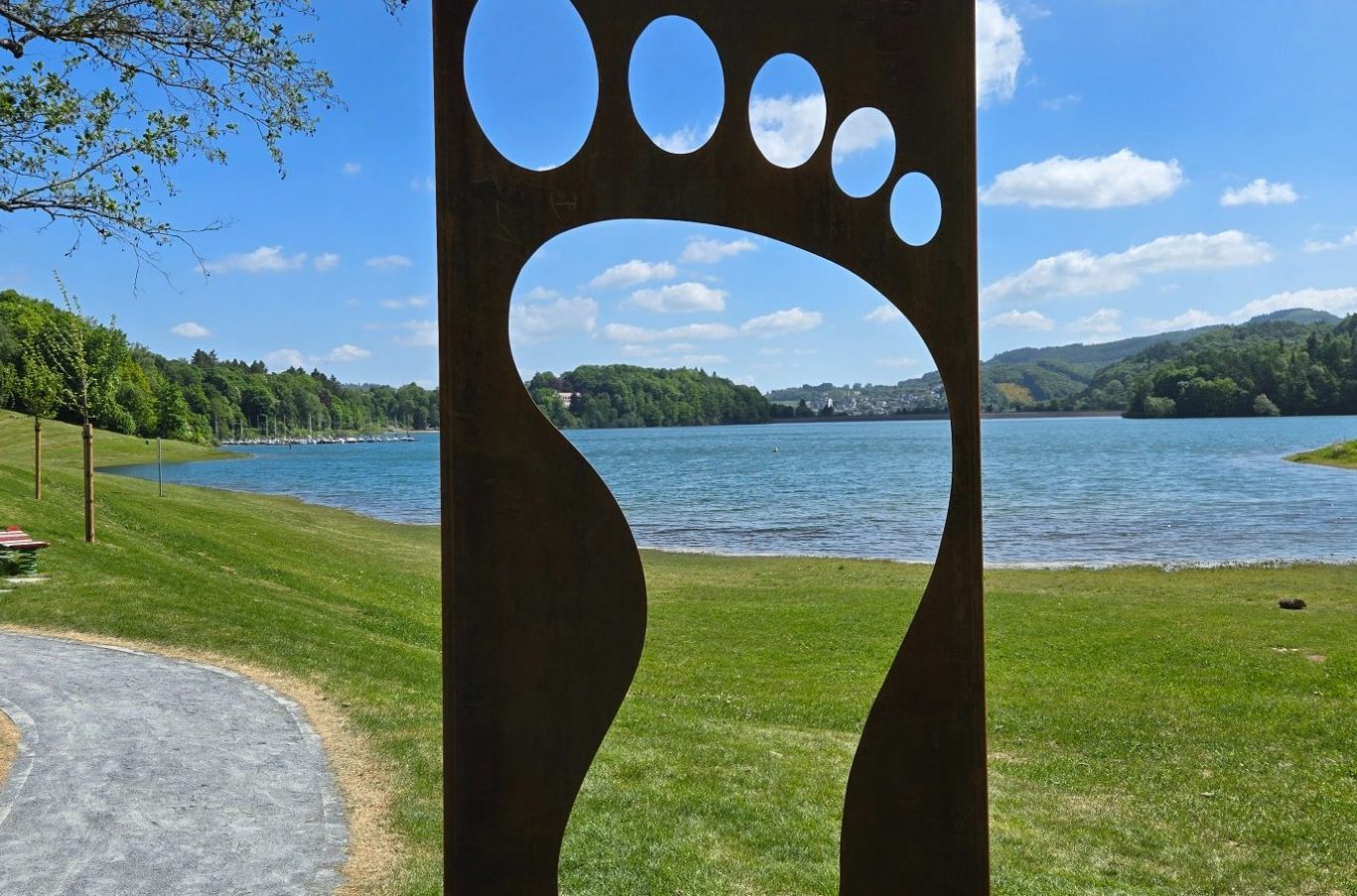 Fußabdruck-Installation vor einem ruhigen See und grünen Hügeln unter blauem Himmel.